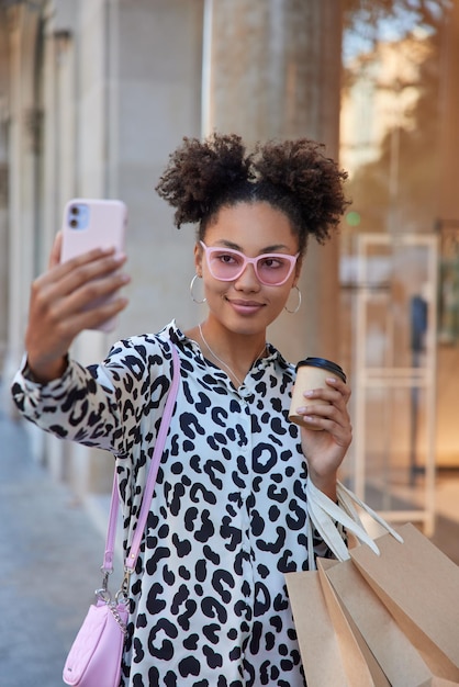 Stylish curly teenage girl returns fro mall with shopping bags drinks takeaway coffee poses for making selfie looks at smartphone camera wears pink sunglasses fashionable shirt stands outside