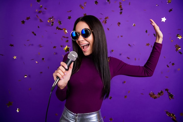 stylish cute woman in eye wear glasses singing into microphone performing her new song isolated vibrant purple color wall