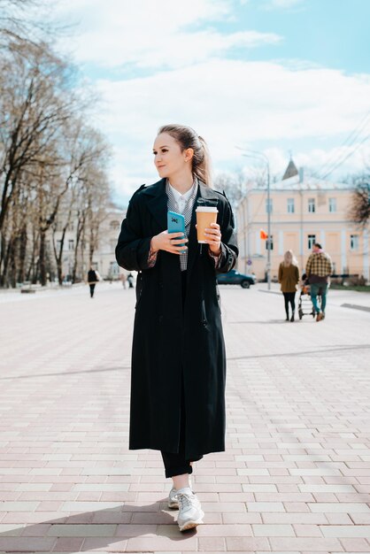 公園を歩きながらスマートフォンとコーヒーを持って黒いコートを着たスタイリッシュなかわいい若い女性