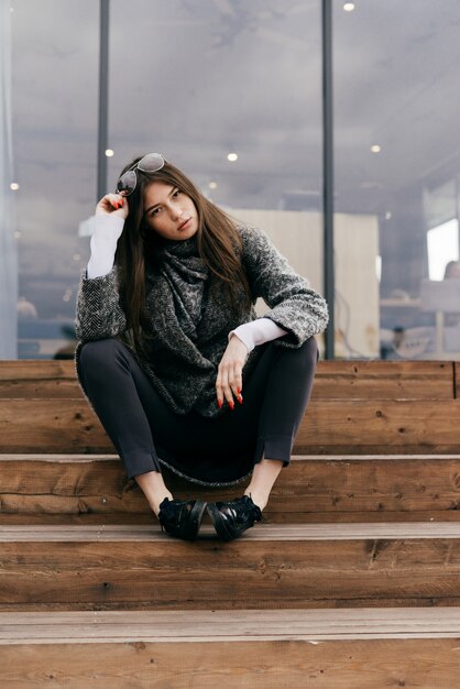 Stylish dark-haired girl in gray coat sits on steps, posing, cloudy weather