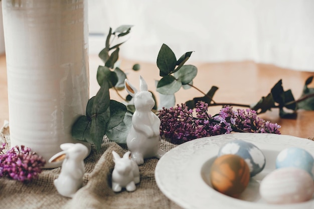 Stylish Easter eggs on vintage plate white bunny rabbits and lilac flowers on rustic fabric on wooden table Rural easter composition of natural dyed eggs and spring purple flowers