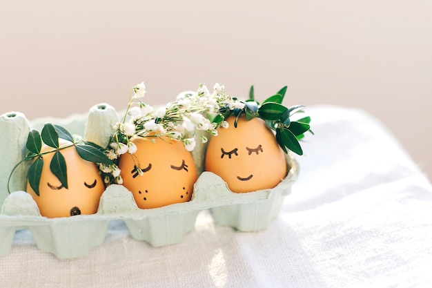 Stylish Easter eggs with cute faces in floral wreath crowns in carton tray on rustic background Modern easter eggs with flowers and sleepy eyes in sunny light Happy Easter eco concept