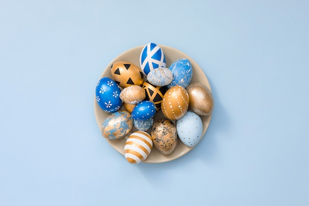 Stylish Easter golden decorated eggs on grey plate isolated on blue surface. Trendy flat lay easter.