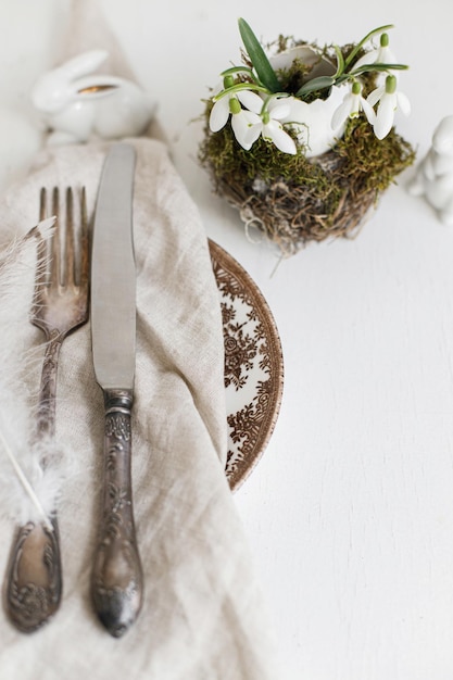 Stylish easter table setting natural egg with snowdrops in nest\
feathers on napkin on modern plate