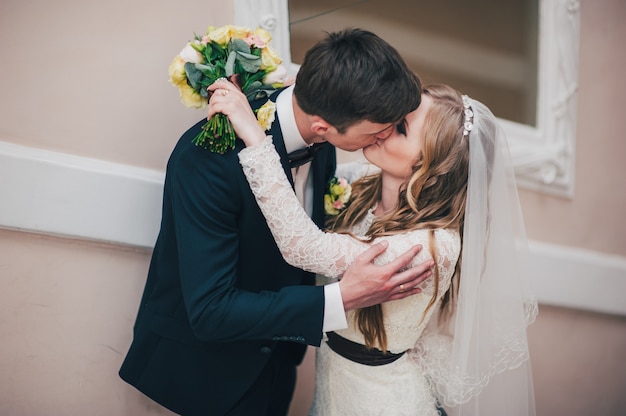 A stylish elegant bride with bouquet of wedding flowers stands near the mirror on the stairs near wall. Kisses the groom in the lips. hugs. Close up. Portrait. Retro. Vintage architecture indoors.