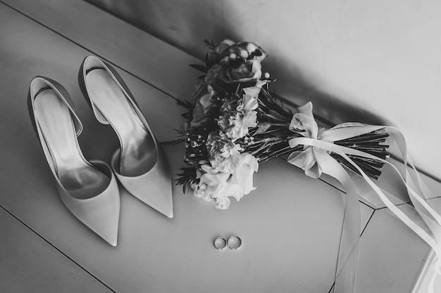 Eleganti scarpe classiche laccate eleganti e bouquet da sposa della sposa di fiori rose e verdi due fedi nuziali d'argento sdraiate sul tavolo primo piano vista laterale foto in bianco e nero