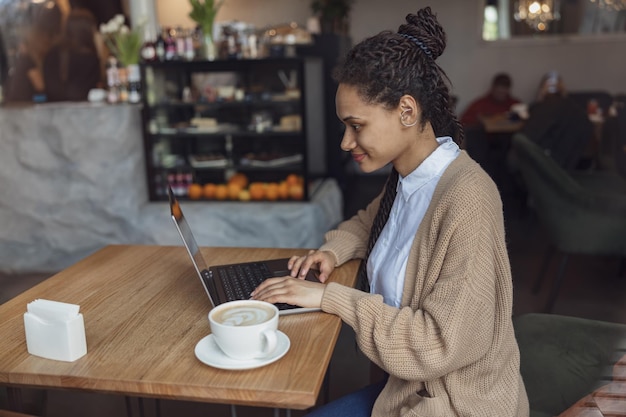 Stylish ethnic student blogger works on laptop in coffee shop Internet online education freelance remote work concept