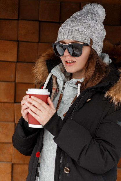 뜨거운 음료와 빨간 컵을 들고 빈티지 나무 벽 근처 선글라스에 모피와 재킷에 따뜻한 운동복에 니트 모자에 세련 된 유럽 젊은 Hipster 여자. 아름다운 현대 소녀.