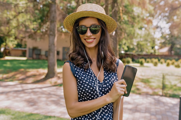 Photo stylish european young smiling woman walking in street in summer dress and hat with coffee and listening music and holding smartphone