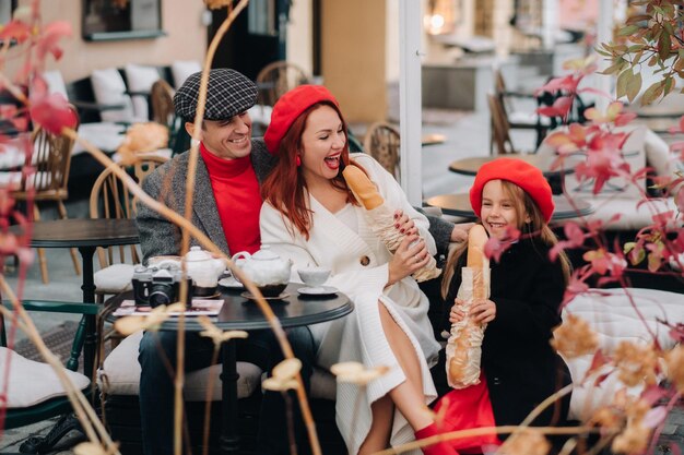 A stylish family of three is sitting at a table outside in a\
cafe and drinking coffee dad mom and daughter in the autumn\
city