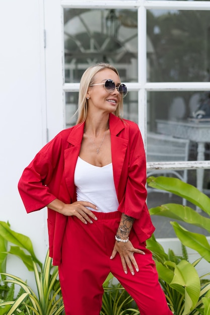 Stylish fashion blonde woman in red suit and white top posing on white wall background tropical leaves natural daylight Wearing trendy sunglasses