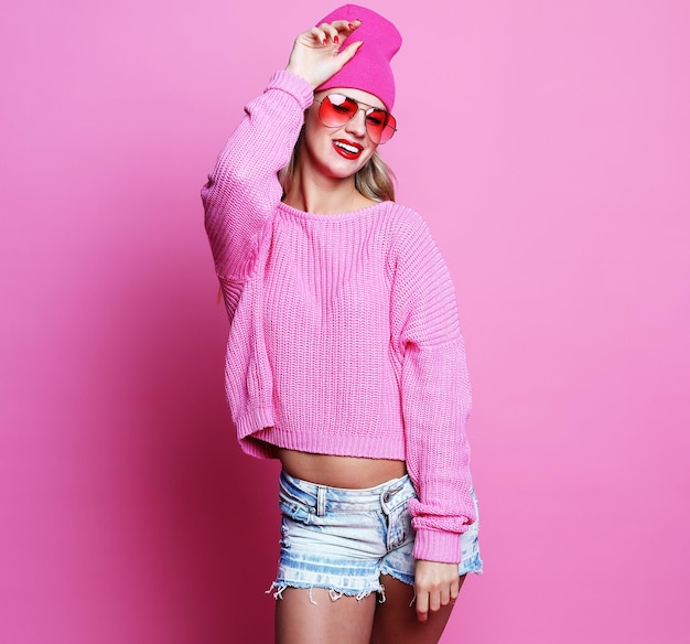 ピンクの背景にポーズをとるピンクのセーターと帽子の流行のカジュアルな若い女性のスタイリッシュなファッションの肖像画ヒップスタースタイル