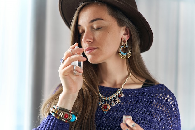 Stylish fashionable attractive brunette boho chic woman with closed eyes wearing jewelry and hat enjoys of perfume scent