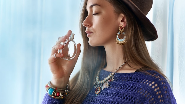 Stylish fashionable attractive brunette boho chic woman with closed eyes wearing jewelry and hat holds perfume bottle