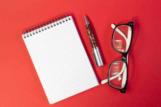 Stylish fashionable glasses in black and white frames notepad and pen on bright red background closeup