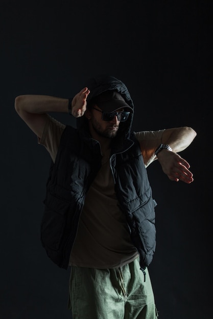 Photo stylish fashionable man dancer with a cap and sunglasses in a hooded vest dances in the dark