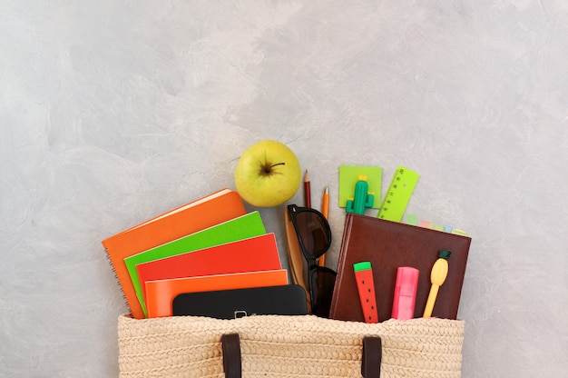 Photo stylish fashionable wicker bag with textbooks and notebooks, green apple, and pens in the form of pineapple, watermelon, cactus, other stationery and sunglasses.