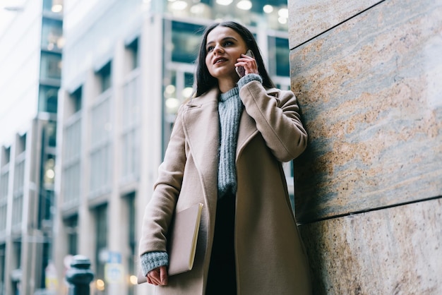 Stylish female in coat with notebook talking on smartphone