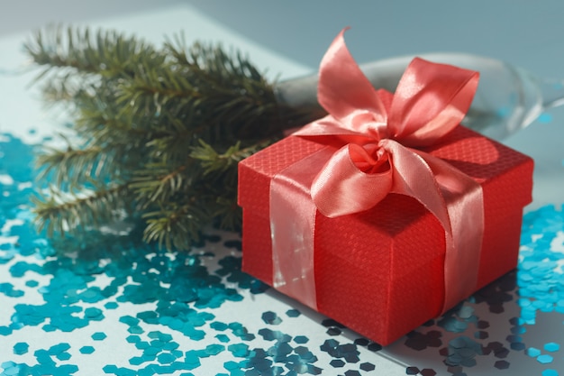 Стильная праздничная композиция с красной подарочной коробкой с атласным коралловым бантом, еловыми ветками и синим посыпанным конфетти.
