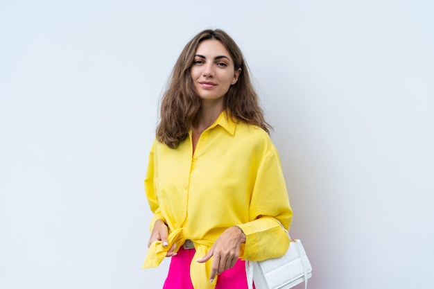 黄色のシャツと明るいピンクのパンツを着たスタイリッシュなファッションの女性が、白い壁の背景に屋外の自然な日の光にトレンディなポーズをとっている