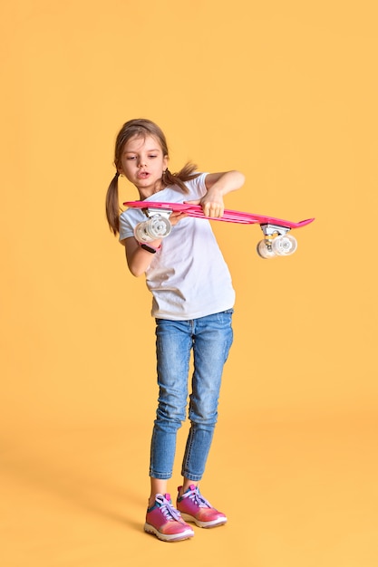 Stylish funny girl wearing white t-shirt, blue jeans and sneakers, holding skateboard over yellow wall