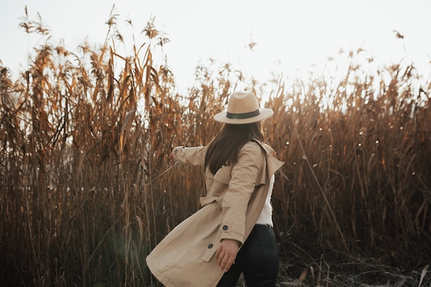 Ragazza alla moda in un cappotto beige e un cappello sullo sfondo di canne.