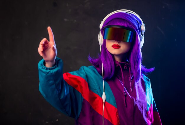 Stylish girl in cyber punk glasses and  tracksuit with headphones on dark wall