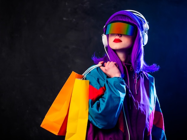 Stylish girl in cyber punk glasses and  tracksuit with shopping bags and headphones on dark wall