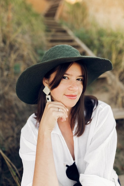 Stylish girl in fashionable look sitting on wooden stairs on beach cliff at sea in sunny light Happy young boho woman in hat relaxing on shore at sunset Summer vacation Fine art