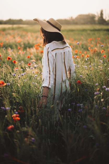 帽子をかぶったおしゃれな女の子が夏の牧草地で夕日の光の中で野生の花の中を歩いている リネンのドレスを着た若い女性が田舎でポピーとヤグルマギクの間を歩いている 田舎のスローライフ シンプルな生活を楽しんでいる