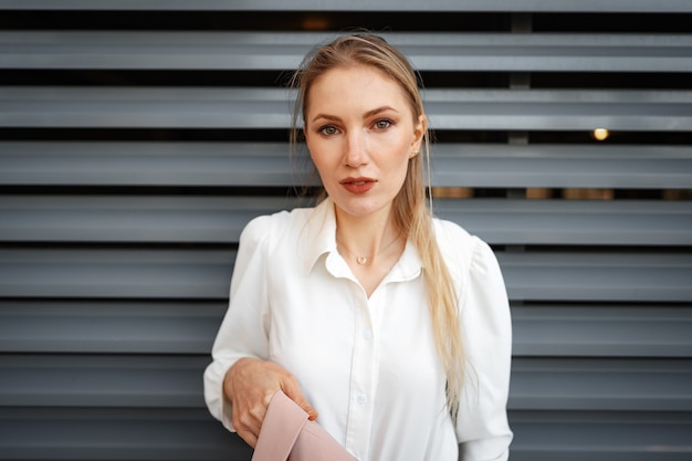 Stylish girl in jeans and white cotton blouse posing near gray wall