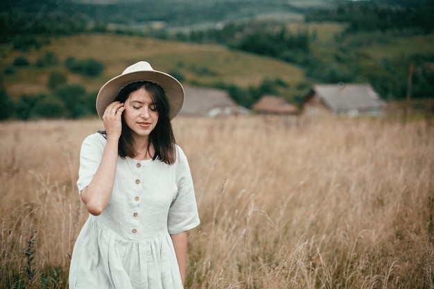 Stylish girl in linen dress and hat walking among herbs and wildflowers in field boho woman smiling and relaxing in countryside simple slow life style space for text atmospheric image