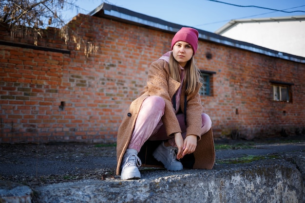 Стильная девушка-модель в коричневом пальто, розовом костюме и серых сапогах на руинах города. Тенденции современной моды. Модный образ