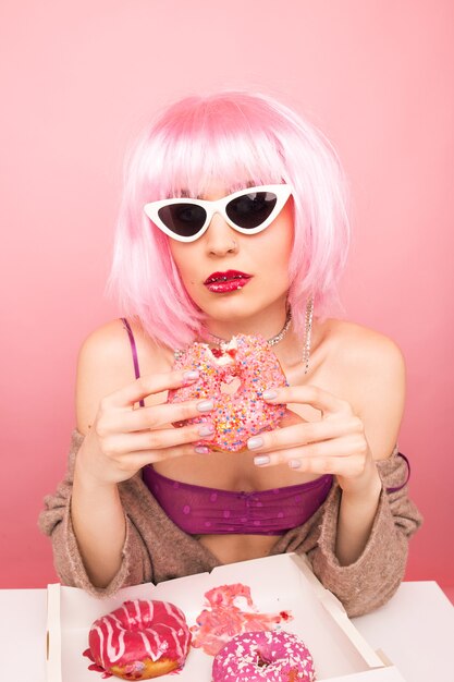 Stylish girl in a pink wig eats delicious donuts on pink