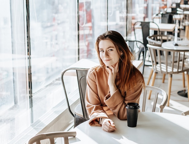 Stylish girl sits in a cafe and drinks coffee. Coffee to go in a cardboard cup. Woman with ginger hair in a beige warm suit in a cozy atmosphere. Modern interior. Calm and pleasant pastime