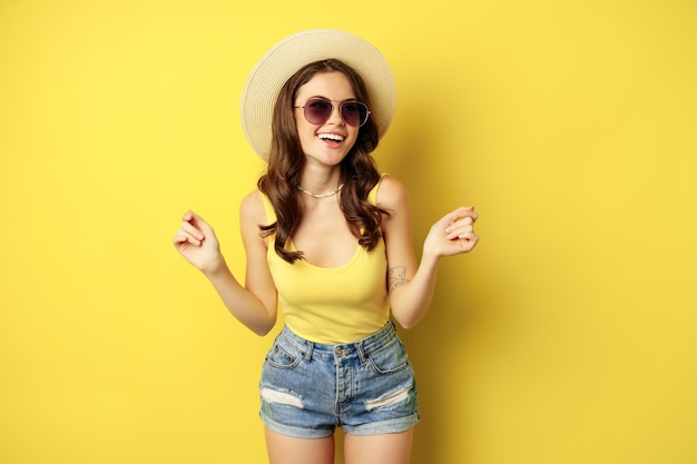 わらの帽子とタンクトップのスタイリッシュな女の子、夏の準備ができて、休暇に行き、黄色の背景の上に立って、喜んで笑っています。