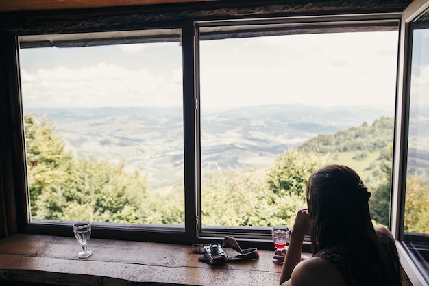 Viaggiatore di ragazza alla moda seduto alla finestra con vista sulle montagne e cielo donna hipster felice che tiene bevanda guardando il cielo e rilassante concetto di vacanza estiva viaggio e voglia di viaggiare