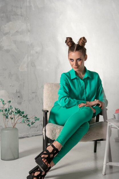 Stylish girl in turquoise shirt and trousers sits elegantly on chair in studio Model with bun hairstyle and evening makeup gray wall background