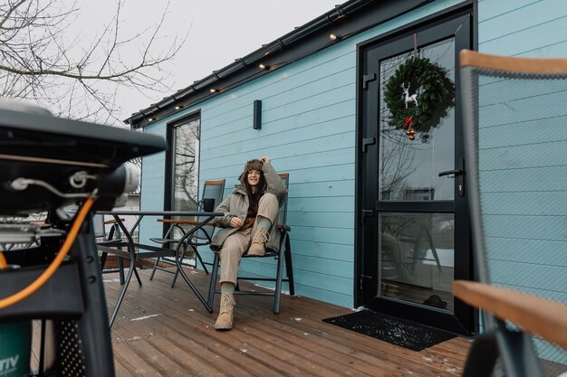 Photo stylish girl in a warm jacket and hat in the yard of a tourist house is grilling meat and drinking coffee enjoying a rural vacation and natural scenery