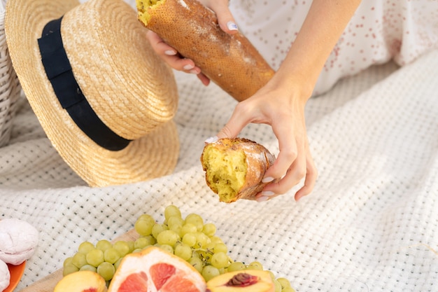 Stylish girl in a white dress breaking a crisp french baguette. The company of cheerful female friends having fun outside in a picnic