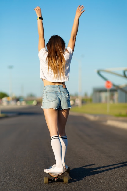 Стильная девушка в белых чулках наслаждается поездкой на лонгборде с поднятыми руками