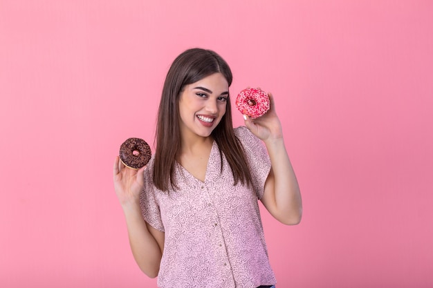 Stylish girl with long hair positively poses, holding fresh pink and chocolate donuts with powder ready to enjoy sweets.