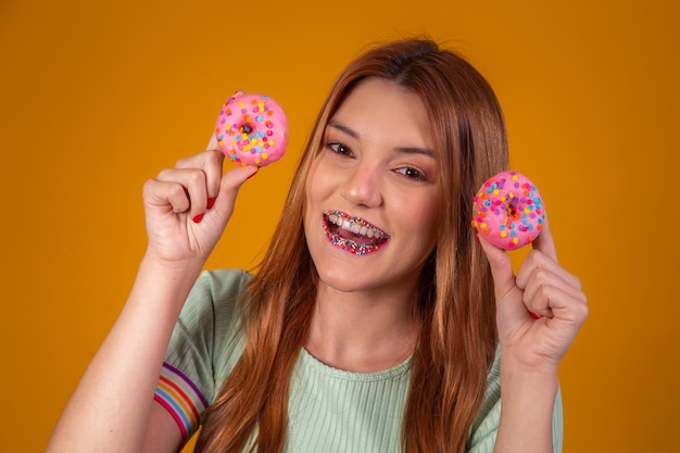 Stylish girl with long hair positively poses, holding fresh pink donuts with powder ready to enjoy sweets.