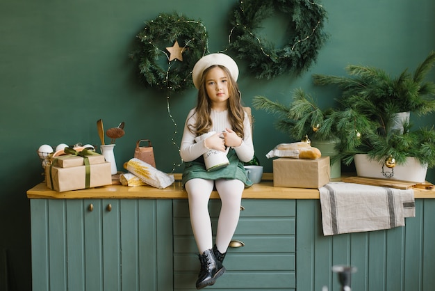 A stylish girl with a pitcher in her hands sits on the kitchen countertop in a room decorated for Christmas and New Year in emerald and green colors.