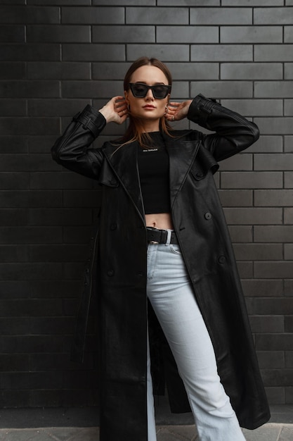 Stylish glamorous young pretty woman model in fashionable black clothes with a fashion long leather coat with a black Tshirt and vintage jeans standing near a black brick wall