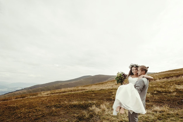 Photo stylish groom carrying happy bride and having fun boho wedding couple luxury ceremony at mountains with amazing view space for text