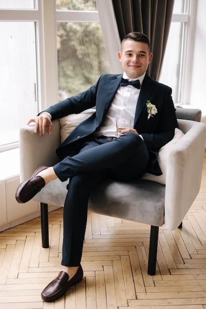 Stylish groom sitting on chair in front of big window man drinks a glass of whiskey wedding day