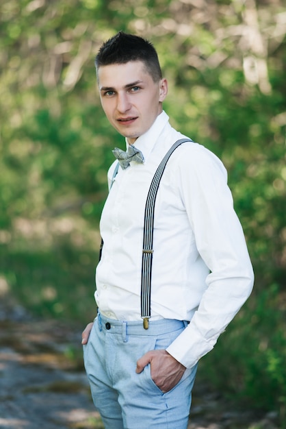 Stylish groom in a white shirt, blue pants, with a bow tie and
suspenders stands