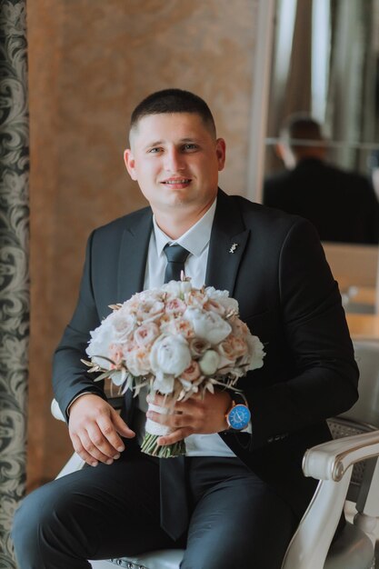Photo a stylish groom with a bouquet of flowers is sitting on a chair in an expensive hotel room grooms morning the groom is getting ready in the morning before the wedding ceremony
