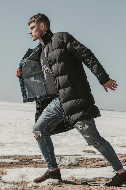 Stylish guy moving forward on the snowy seashore in a black down coat and scruffy jeans.